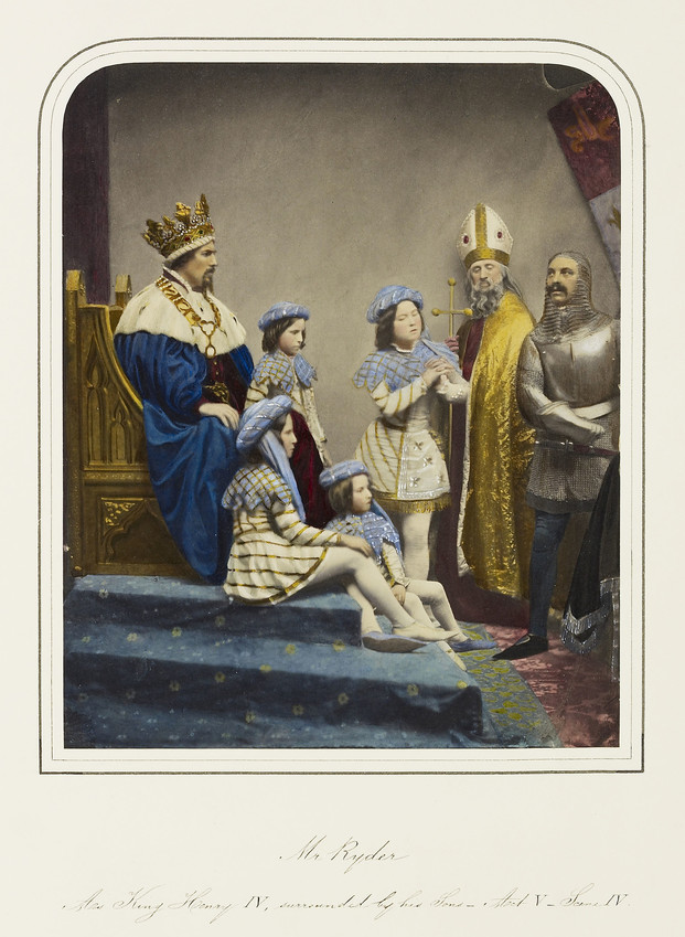A photograph, hand-coloured in watercolour, of a group of actors performing in Richard II. On the left, in profile, John Ryder as Bolingbroke is seated on a gold-painted throne on a carpeted pedestal. He wears a royal blue cloak with an ermine collar over a red tunic, a gold chain of office, and an elaborate gold crown. He is surrounded by four boys: two sit at his feet, the other two stand beside and in front of him. All four children wear matching outfits: a cream tunic with gold embroidery, a pale blue collar with a zig-zag hem, a blue hat, white stockings, and one blue and one white shoe. In the background stands a bishop in mitre and gold robe, holding a crosier. On the right stands a soldier in plate armour and a chainmail hood.