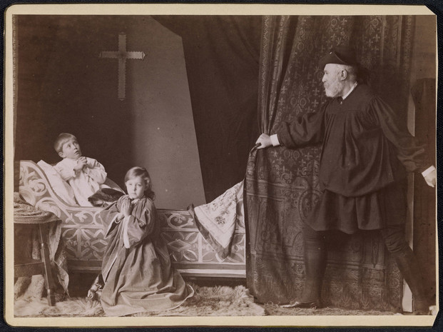 A photograph of an adult man and two children posing in a scene from Richard III. A small boy sits on a chaise-longue in a white night-shirt, his hands clasped in prayer and his gaze directed heavenward. On the floor in the foreground, a girl, slightly older, kneels on a fur rug; she wears a cloak with voluminous sleeves, and is also praying, but her gaze is directed outwards toward the viewer. On the right, a white bearded man in black tunic, boots, and hat is pulling back a brocade curtain. There is a cross on the back wall.