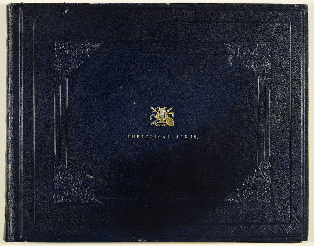 A large album, landscape format, bound in dark blue leather. Patterns are inlaid into the leather on the front cover, framing the gold embossed lettering saying ‘Theatrical Album’ and an emblem made up from a harp, theatrical mask, sword, arrow, and chain.
