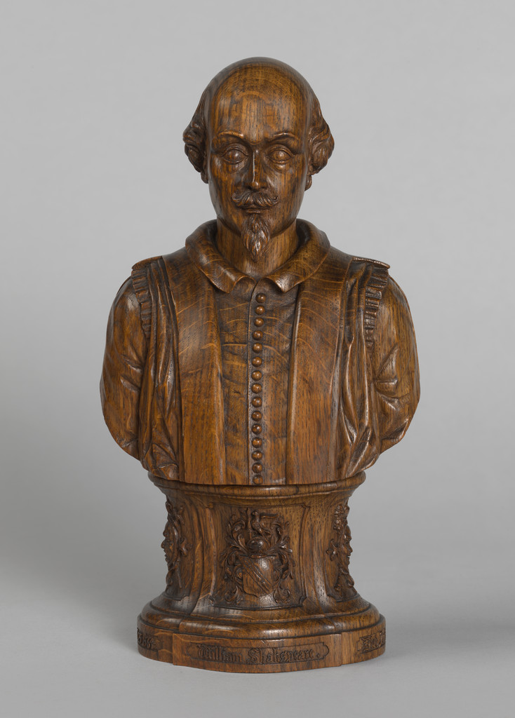 A polished wooden bust of Shakespeare on a small plinth. "William Shakespeare", "Born 1564" and "Died 1616" are carved around the base in a blackletter style. Shakespeare's coat of arms and the masks of comedy and tragedy are carved on the plinth. Shakespeare is recognisable by his bald head, short beard, and costume with a narrow collar and row of small buttons, based on that of his funerary monument in Statford.