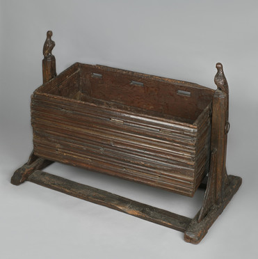 A dark wooden cradle, rectangular, mounted on a stand to allow it to be rocked. The stand is decorated with carved birds at each end. The cradle is carved in horizontal creases, and has three narrow holes near its upper edge on either side. Both are visibly battered with age.
