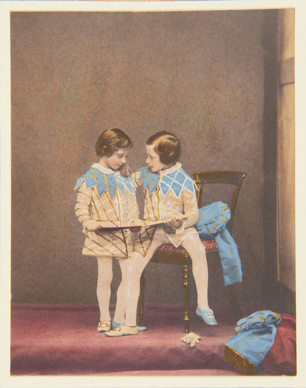 A photograph, hand-coloured in watercolour, of two children in medieval fancy dress: both wear short cream tunics with gold embroidery and pale blue collars with zig-zag hems, white stockings, and one blue and one white shoe. The taller child is perched on the edge of a Victorian wooden chair with an upholstered seat. He has his hand on the smaller child’s shoulder, looking at him, as they both hold an opened folder or slim volume. Two blue fabric hats, matching the collars, have been discarded on the chair and the floor; a white glove is on the floor by the chair.